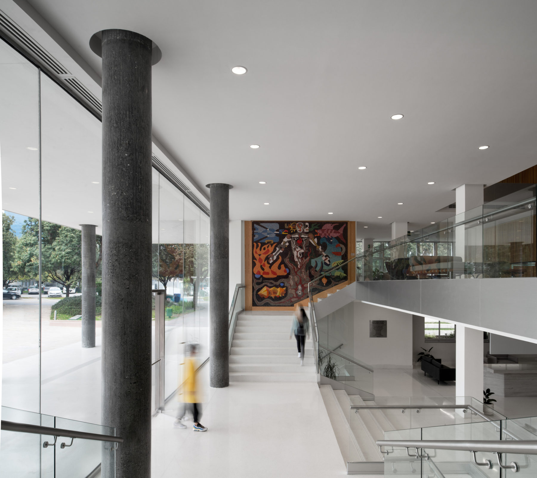 Interior photo of building entry from a side view. One person enters, another is walking up stairs toward mural.