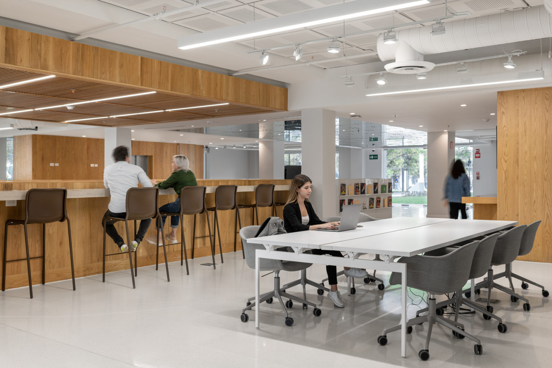 Interior photo of building. One student sits at a white table on a laptop. Two others sit at bar height chairs in conversation.